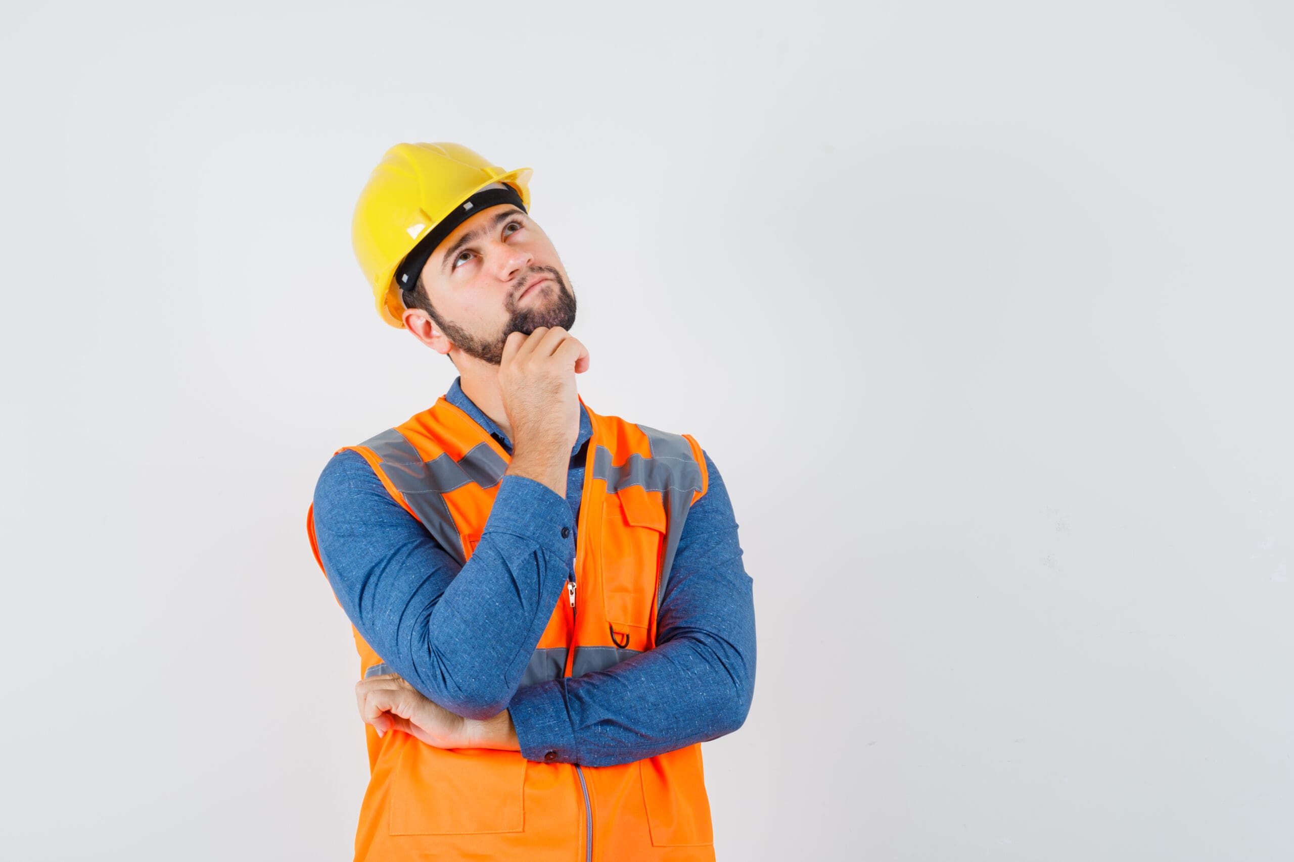 Joven constructor mirando hacia arriba con camisa, chaleco, casco y mirando pensativo