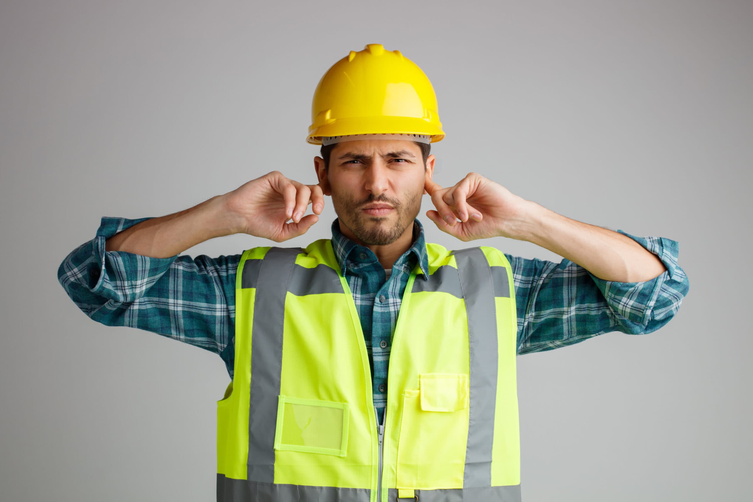 Ingeniero joven descontento con casco de seguridad y uniforme mirando a la cámara mientras mantiene los dedos en los oídos aislados sobre fondo blanco