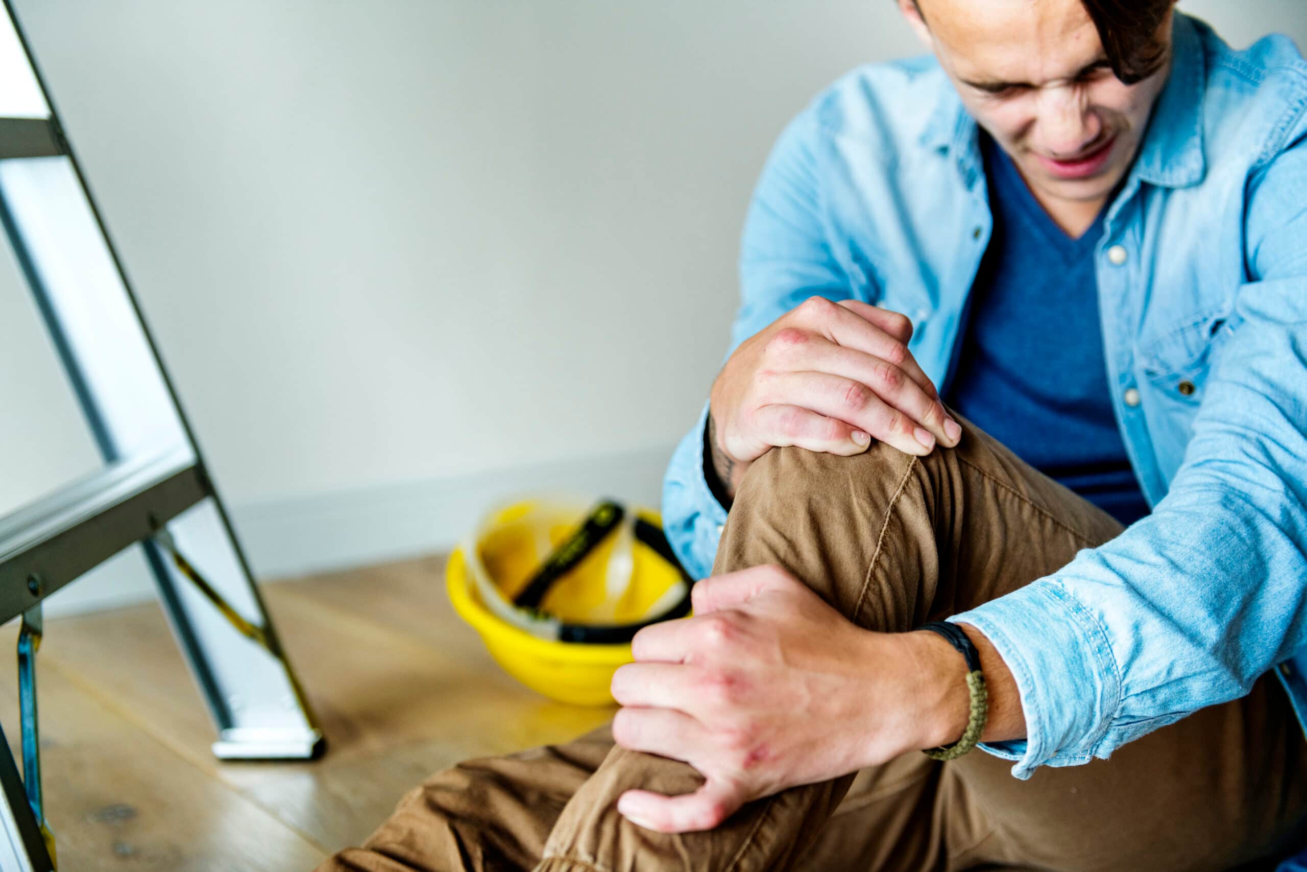 Un hombre se sienta en el suelo sujetándose la rodilla lesionada, con aspecto de dolor, con un casco y una escalera cerca, lo que sugiere un accidente laboral.